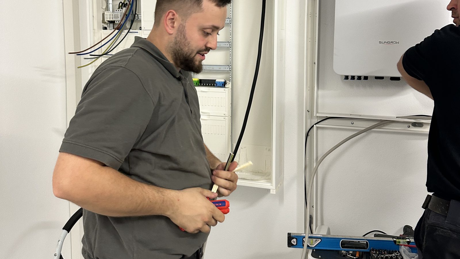 Monteur am geöffneten Zählerschrank, rechts daneben Wechselrichter an der Wand und Batteriespeicher auf dem Boden